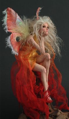 a woman dressed as a fairy sitting on top of a tree stump with red and yellow feathers