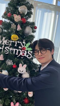 a man holding up a merry christmas sign next to a christmas tree with ornaments on it