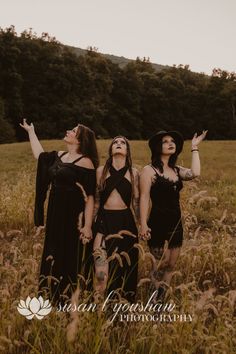 three women standing in tall grass with their hands up and one woman wearing a black dress