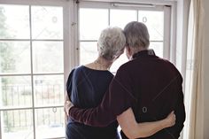 an older couple standing in front of a window with their arms around each other and looking out the window