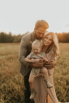 a man and woman holding a baby in an open field with the sun behind them