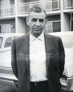 black and white photograph of an older man standing in front of a building with cars behind him