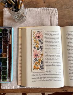 an open book sitting on top of a wooden table next to watercolor paints and brushes