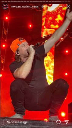 a man in black shirt and orange hat on stage with his hands up to the air