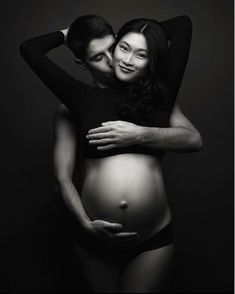 a pregnant woman is hugging her husband's head while they are posing for a black and white photo