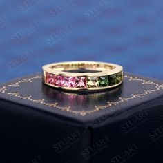 a yellow gold ring with pink, green and blue stones on it sitting on top of a black box