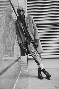 a man leaning against a wall wearing a coat and hat