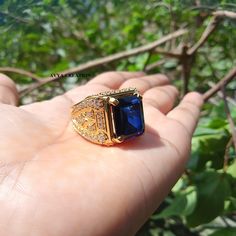 a hand holding a ring with a blue stone in it's center, surrounded by greenery