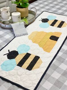 a quilted table runner with two bees on it and a potted plant in the background