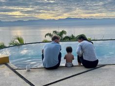 two adults and a child sitting on the edge of a swimming pool with an ocean view