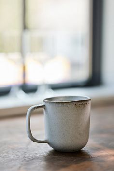 a white coffee cup sitting on top of a wooden table next to a large window