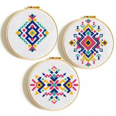 three cross stitch hoops with colorful designs on white fabric and gold trimming, each featuring an embroidered design