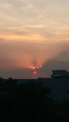 the sun is setting behind some clouds in the sky over a building and tree line