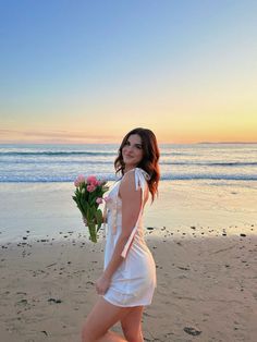 a woman is walking on the beach with flowers in her hand and she is wearing a white dress