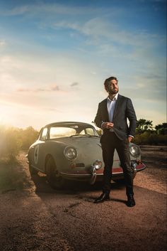 a man in a suit standing next to a car on a dirt road with the sun behind him