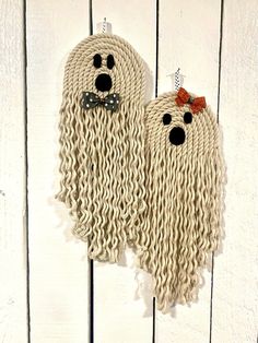 two knitted ghost decorations hanging on a white wooden wall, one with a bow tie and the other with eyes