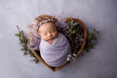 a newborn baby wrapped in a purple wrap is laying on a basket with flowers and greenery