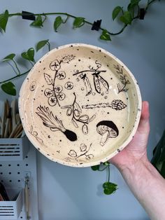 a hand holding a bowl with drawings on it and plants growing in the back ground