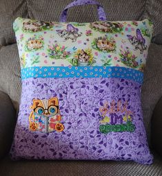 a purple pillow with cats and flowers on it sitting on a couch next to a gray chair