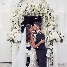 a bride and groom kissing in front of a white arch with flowers on the side