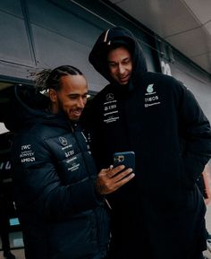 two men standing next to each other in front of a building looking at a cell phone