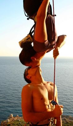 two men hanging upside down on the side of a cliff by the ocean with their hands in the air