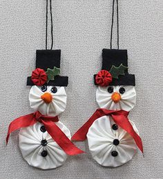 two snowman ornaments hanging from strings on a white wall with red ribbon and holly