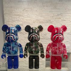 three different colored teddy bears on display in front of a white brick wall, one with a shark's mouth