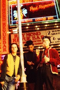 three young men standing on the sidewalk in front of a neon sign at night time