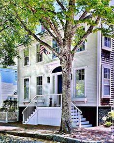 a white house sitting next to a tree on the side of a road with stairs leading up to it