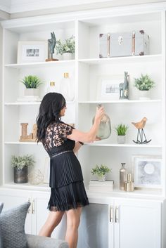 a woman standing in front of a white book shelf filled with plants and vases