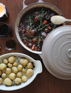 potatoes and meat in a pot next to wine glasses on a wooden table with utensils