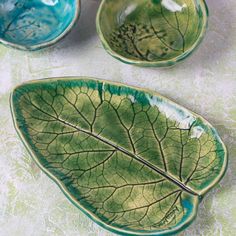 three green leaf shaped dishes sitting on top of a white tableclothed surface with leaves painted on them