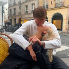 a man and woman sitting on a bench in front of a building kissing each other