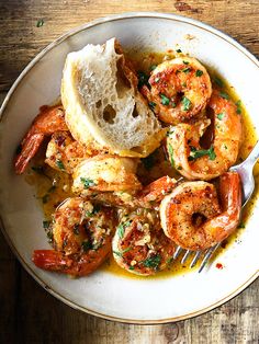 a white bowl filled with shrimp and bread on top of a wooden table next to a fork