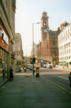 people are walking on the sidewalk in front of buildings and a clock tower behind them