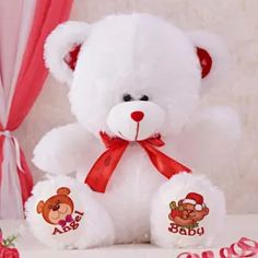 a white teddy bear with red ribbon around its neck and name on the chest sitting in front of pink curtains