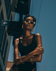 a woman standing next to a building with her arms crossed wearing sunglasses and a black dress