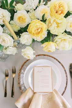 a table setting with white and yellow flowers in the background, gold rimmed place settings