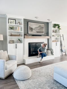 a woman sitting on the edge of a fireplace in a living room with white furniture