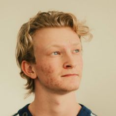 a young man with freckled hair and blue eyes looks into the distance while standing in front of a white wall