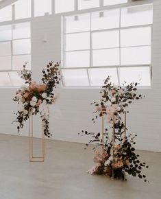 two tall metal vases with flowers and greenery on them in front of a window