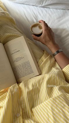 a person laying in bed reading a book and holding a cup of coffee with both hands