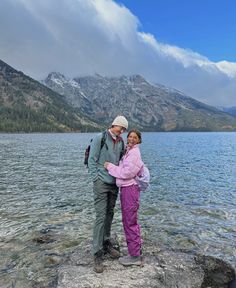 a man and woman standing next to each other on top of a rock near the water