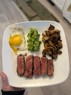 a person holding up a plate with meat, mushrooms and an egg on the side