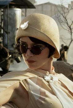 a woman wearing a hat and sunglasses sitting in a chair with other people behind her