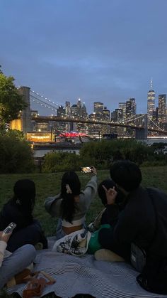 four people sitting on a blanket in front of the city skyline at night, taking pictures with their cell phones