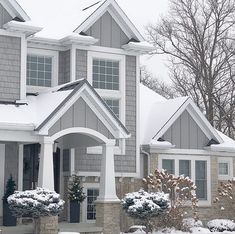 a house with snow on the ground and bushes in front of it, surrounded by trees