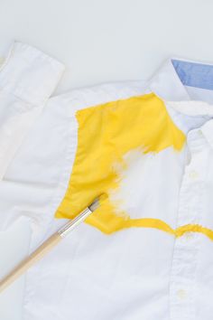 a white shirt with yellow paint on it and a brush next to it, all in the same color