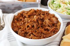 pulled pork in a white bowl next to coleslaw and bread on a table